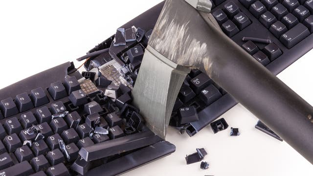 Eine schwarze Computertastatur, die mit einer Axt in der Mitte zerschlagen wurde. Tasten und Teile der Tastatur sind zerbrochen und verstreut. Die Axt ist in einem Winkel in die Tastatur eingedrungen, was auf eine gewaltsame Handlung hindeutet. Die Szene ist auf einem weißen Hintergrund dargestellt.