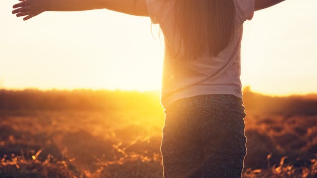 Ein junges Mädchen steht mit ausgebreiteten Armen in einem Feld bei Sonnenuntergang. Die Sonne strahlt warmes, goldenes Licht aus, das die Szene erhellt. Die Atmosphäre wirkt friedlich und frei.