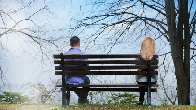 Zwei Personen sitzen weit entfernt voneinander auf einer Parkbank. Sie sind nur von hinten zu sehen: links eine Person mit kurzen dunklen Haaren, rechts eine Person mit langen blonden Haaren. Die kahlen Bäume und der wolkige Himmel verleihen der Szene eine ruhige und etwas traurige Atmosphäre.