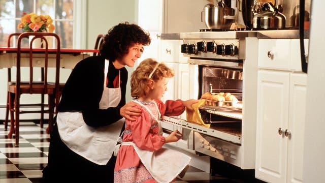 Eine Frau und ein Kind, beide in Schürzen, stehen in einer Küche vor einem geöffneten Backofen. Frisuren und Kleidung der beiden lassen darauf schließen, dass die Aufnahme bereits etwas älter ist. Die erwachsene Frau legt dem Kind sanft die Hand auf die Schulter, während es ein Backblech mit Gebäck in den Ofen schiebt. Im Hintergrund ist ein Tisch mit Blumen zu sehen. Die Szene vermittelt eine warme, häusliche Atmosphäre.