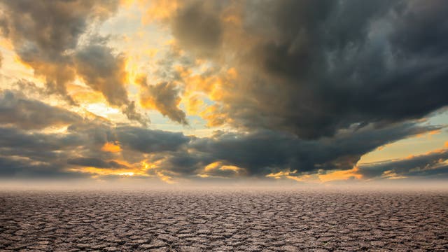 Weite, trockene Landschaft mit rissigem Boden unter einem dramatischen Himmel. Dunkle Wolken ziehen auf, während die untergehende Sonne den Himmel in warmen Gelb- und Orangetönen färbt. Die Szene vermittelt ein Gefühl von Trockenheit und Wetterveränderung.