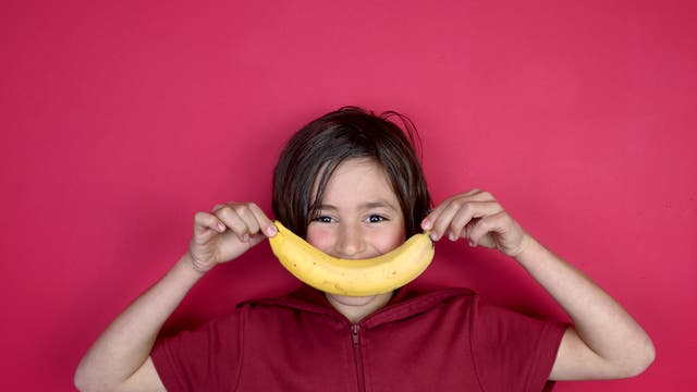 Ein Junge vor pinkfarbenem Hintergrund hält eine Banane vor seinen Mund, so dass sie sein Lächeln ergibt. 