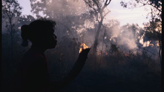 Eine Angehörige eines indigenen Volkes in Australien hält einen brennenden Ast. In Hintergrund lodern Flammen im Unterholz.