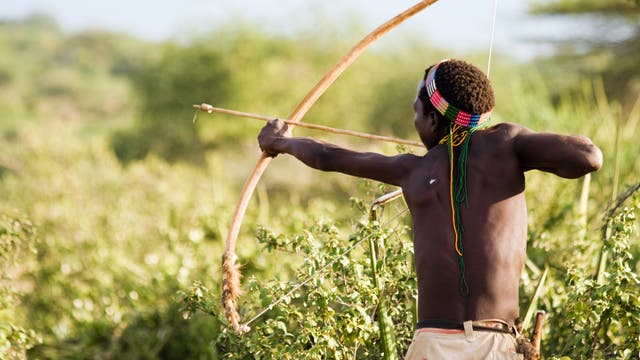 Eine Person steht in einer natürlichen Umgebung und spannt einen Bogen, bereit, einen Pfeil abzuschießen. Sie trägt ein buntes Stirnband und hat den Blick auf das Ziel gerichtet. Im Hintergrund ist dichte Vegetation zu sehen, die eine ländliche oder wilde Landschaft andeutet.