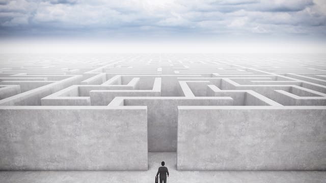 Eine Person in einem Anzug steht vor einem großen, komplexen Labyrinth aus hohen, grauen Wänden, das sich bis zum Horizont erstreckt. Der Himmel ist bewölkt, was eine nachdenkliche und herausfordernde Atmosphäre schafft. Die Person hält einen Aktenkoffer und scheint bereit, das Labyrinth zu betreten, was symbolisch für eine bevorstehende Herausforderung oder Entscheidung steht.