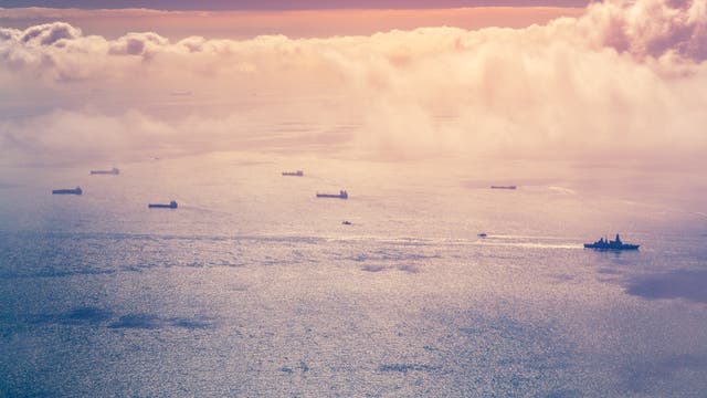 Mehrere Frachtschiffe fahren auf einem ruhigen Meer unter einem Himmel mit rosa und orangefarbenen Wolken. Die Sonne spiegelt sich auf der Wasseroberfläche und erzeugt ein glitzerndes Muster. Die Szene vermittelt Ruhe und Weite.