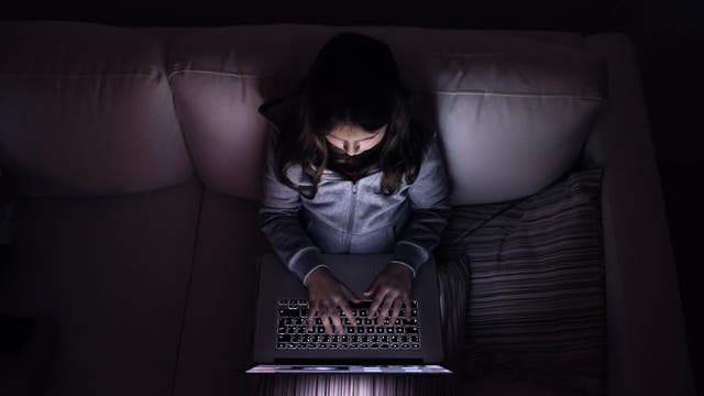 Ein Mädchen sitzt in einem dunklen Raum auf einem Sofa und tippt auf einem Laptop. Die Szene vermittelt eine düstere, einsame Atmosphäre.