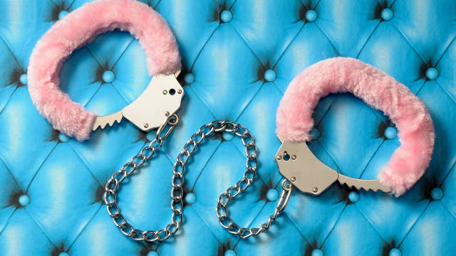 Ein Paar Handschellen mit rosa Plüschüberzug liegt auf einer blauen, gesteppten Oberfläche. Die Handschellen sind durch eine silberne Kette verbunden. Die Szene vermittelt einen Kontrast zwischen der weichen Textur des Plüschs und der metallischen Härte der Handschellen.