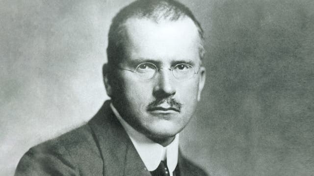 Ein Schwarz-Weiß-Foto eines Mannes mit Brille und Schnurrbart, der direkt in die Kamera schaut. Der Hintergrund ist neutral und unscharf, wodurch der Fokus auf das Gesicht des Mannes gelegt wird.