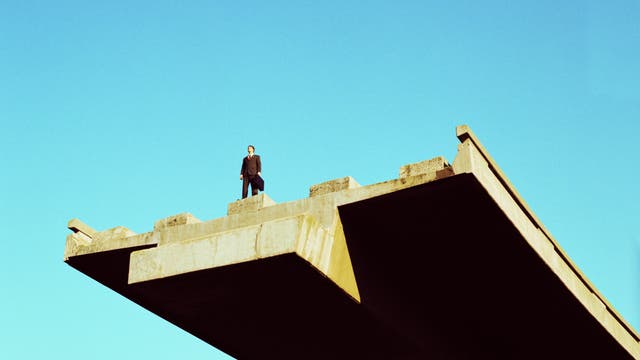 Eine Person in dunklem Mantel steht an der Kante eines unvollendeten Brückenabschnitts. Der Himmel ist klar und blau. Die Brücke aus Beton endet abrupt, was den Eindruck von Unvollständigkeit und offener Zukunft vermittelt.