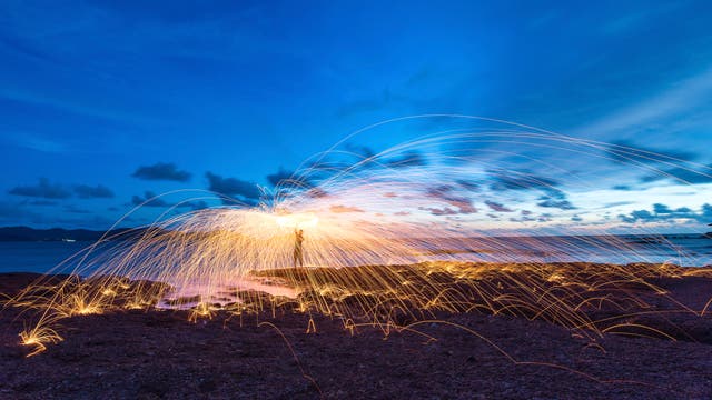 Eine Person steht auf einem felsigen Strand bei Sonnenuntergang und schwingt brennende Stahlwolle, wodurch Funken in einem weiten Bogen sprühen. Der Himmel ist in tiefem Blau mit vereinzelten Wolken gefärbt, und das Meer ist im Hintergrund sichtbar. Die Funken erzeugen leuchtende, geschwungene Linien, die sich über den Boden erstrecken.
