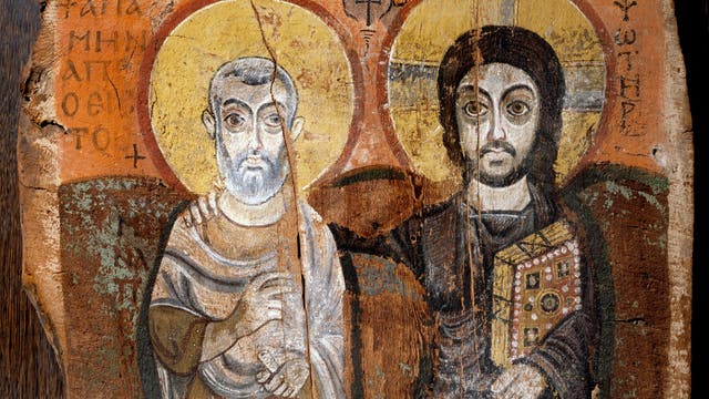 Ein antikes Gemälde auf Holz zeigt zwei heilige Figuren mit Heiligenscheinen. Die linke Figur trägt ein langes Gewand und hält eine Schriftrolle, während die rechte Figur ein reichverziertes Buch hält. Beide Figuren sind frontal dargestellt und von griechischem Text umgeben. Der Hintergrund ist in warmen Erdtönen gehalten, was dem Bild eine historische und religiöse Atmosphäre verleiht.