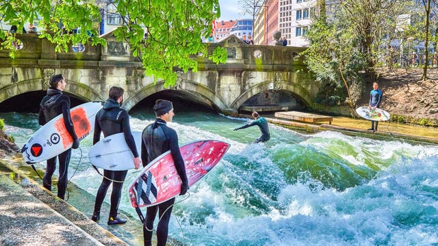 Surfer in Neoprenanzügen stehen mit ihren Surfbrettern am Ufer eines Flusses, während ein Surfer auf einer stehenden Welle reitet. Im Hintergrund ist eine Steinbrücke mit Bögen zu sehen, und Zuschauer beobachten das Geschehen. Die Szene spielt sich in einer urbanen Umgebung mit Gebäuden und Bäumen ab.