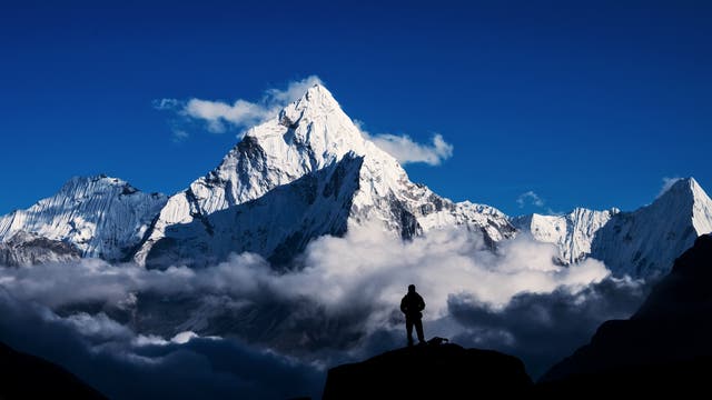 Der Mount Everest ragt weißgrau und schroff in den blauen Himmel auf, eine kleine weiße Wolke befindet sich hinter seinem Gipfel. Im Vordergrund schweben ebenfalls kleinere weiße Wolken in tieferen Lagen. Die schwarze Silhouette eines Mannes steht auf einem schwaren Felsen im Vordergrund