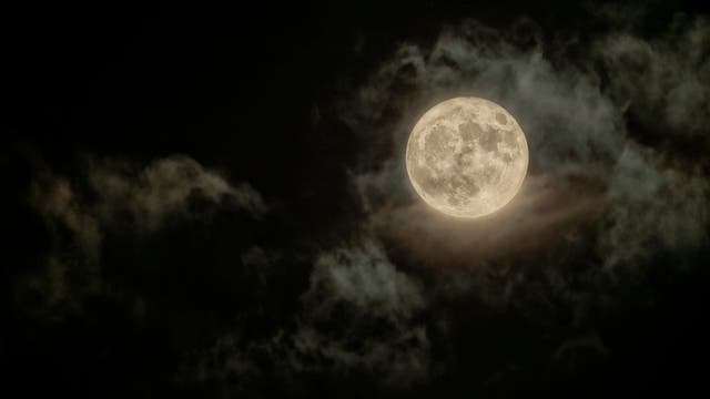 Ein Vollmond leuchtet hell am Nachthimmel, umgeben von dünnen, dunklen Wolken, die ihm einen mystischen Rahmen verleihen. Die Oberfläche des Mondes zeigt deutliche Krater und Strukturen, die typisch für seine Erscheinung sind. Der Kontrast zwischen dem hellen Mond und dem dunklen Himmel betont die Details der Mondoberfläche.