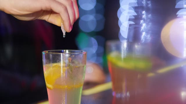 Eine Hand drückt einen Tropfen aus einem kleinen Fläschchen in ein Glas mit einem Getränk, das eine Zitronenscheibe enthält. Im Hintergrund sind verschwommene Lichter zu sehen, die eine lebendige und festliche Atmosphäre schaffen. Das Bild vermittelt den Eindruck einer Bar oder eines Partyumfelds.