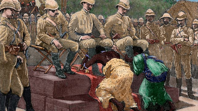 Eine historische Illustration zeigt eine koloniale Szene, in der mehrere uniformierte Männer auf erhöhten Stühlen sitzen. Vor ihnen knien zwei Personen mit gesenkten Köpfen in unterwürfiger Haltung auf einem roten Teppich und umfassen die Beine der Männer. Sie tragen farbenfrohe, traditionell wirkende  Kleidung. Im Hintergrund stehen weitere uniformierte Männer in einer Reihe. Die Szene vermittelt ein Gefühl von Hierarchie und Machtungleichheit.