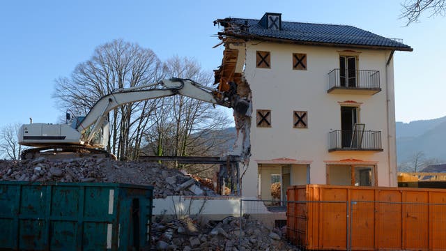 Ein Bagger reißt ein mehrstöckiges Gebäude ab, wobei die Hälfte des Gebäudes bereits zerstört ist. Im Vordergrund sind Trümmer und zwei große Container, einer grün und einer orange. Im Hintergrund sind kahle Bäume und Hügel unter einem klaren blauen Himmel zu sehen.