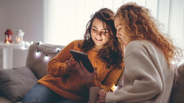 Mutter und jugendliche Tochter sitzen auf einem Sofa und blicken gemeinsam auf ein Tablet.