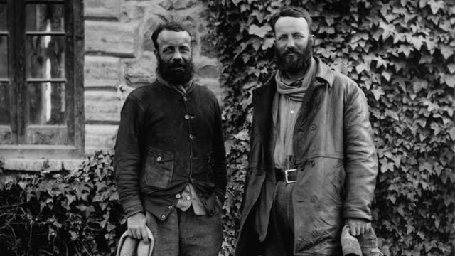 Zwei Männer stehen vor einer mit Efeu bewachsenen Steinwand. Beide tragen wetterfeste Kleidung und haben Bärte. Links im Bild ist ein Fenster zu sehen. Die Szene wirkt historisch und rustikal.