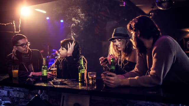 Eine Gruppe von vier Personen sitzt an einer Bar in einem Klub oder einer Lounge. Die Szene ist in gedämpftem Licht gehalten, mit einer warmen, gemütlichen Atmosphäre. Zwei Personen unterhalten sich, während eine andere auf ihr Handy schaut und lächelt. Auf der Theke stehen Getränke, darunter Bierflaschen und Gläser. Im Hintergrund sind bunte Lichter und eine Diskokugel zu sehen, die auf eine entspannte, gesellige Umgebung hinweisen.