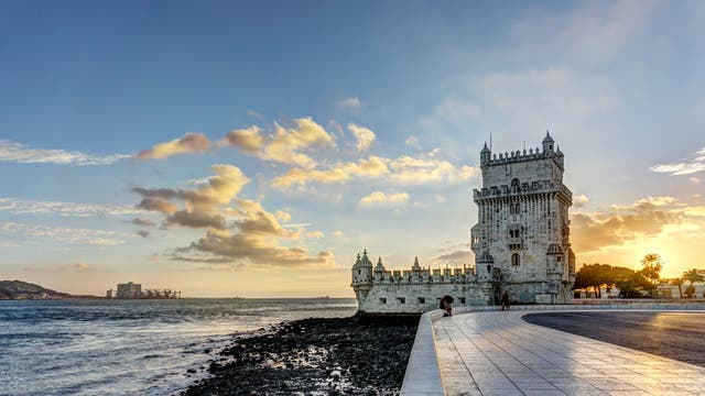 Ein historischer Turm mit Zinnen, Erkern und Türmchen am Ufer eines Flusses bei Sonnenuntergang. Der Himmel ist teils bewölkt, und das Licht der untergehenden Sonne wird auf dem Wasser und den Steinen am Ufer reflektiert. Eine Promenade führt am Wasser entlang, im Hintergrund sind Hügel und Gebäude zu sehen.