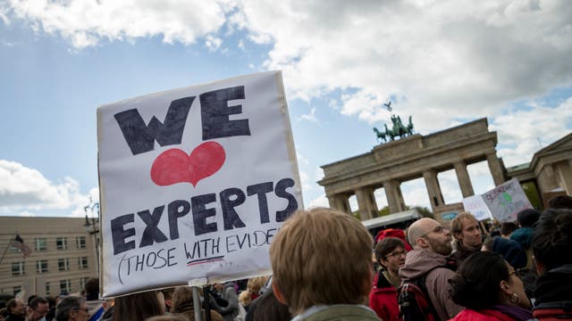 Eine Menschenmenge versammelt sich vor dem Brandenburger Tor. Im Vordergrund hält jemand ein Schild mit der Aufschrift "We ♥ Experts (Those with Evidence)". Der Himmel ist bewölkt, und die Szene vermittelt eine Atmosphäre des Protests bzw. der Unterstützung für wissenschaftliche Expertise.