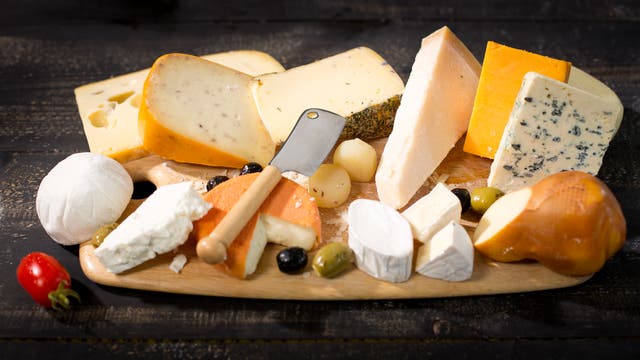 Eine Auswahl verschiedener Käsesorten auf einem Holzbrett, darunter Brie, Blauschimmelkäse, Cheddar und Ziegenkäse. Ein kleines Messer liegt auf dem Brett, umgeben von Oliven, Trauben und einer Kirschtomate. Die Szene ist auf einem dunklen Holztisch arrangiert.