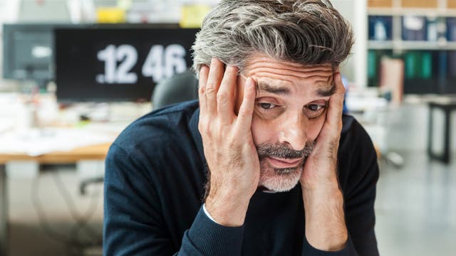 Ein Mann sitzt an einem Schreibtisch und stützt seinen Kopf in die Hände, wirkt nachdenklich oder gestresst. Im Hintergrund ist ein unscharfer Bildschirm mit der Uhrzeit 12:46 zu sehen. Der Raum ist hell beleuchtet und es sind unscharfe Bürogegenstände im Hintergrund erkennbar.