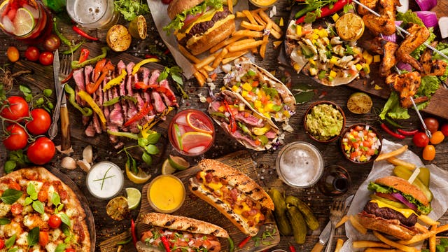 Eine reichhaltige Auswahl an verschiedenen Gerichten auf einem rustikalen Holztisch. Zu sehen sind ein Burger mit Pommes, gegrillte Fleischspieße, Tacos mit buntem Gemüse, ein Steak mit Paprika, eine Pizza mit Tomaten und Basilikum, Hot Dogs mit Senf und Ketchup sowie verschiedene Dips und Saucen. Frische Zutaten wie Tomaten, Zwiebeln und Kräuter sind ebenfalls verteilt. Getränke wie Bier und Cocktails ergänzen das Arrangement. Keine Personen sind im Bild zu sehen.