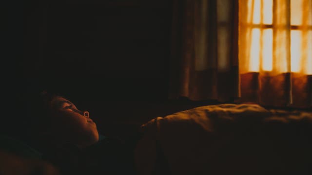 Ein Kind liegt in einem abgedunkelten Raum, während etwas Licht durch ein Fenster mit Vorhängen scheint. Sein Gesicht ist dem Fenster zugewandt.