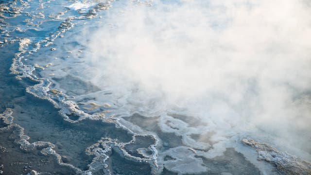 Dampf steigt von einer geothermischen Quelle auf, die von mineralischen Ablagerungen umgeben ist. Die Oberfläche zeigt komplexe Muster aus weißen und grauen Ablagerungen, die sich über das Wasser erstrecken. Die Szene vermittelt eine ruhige, natürliche Atmosphäre.