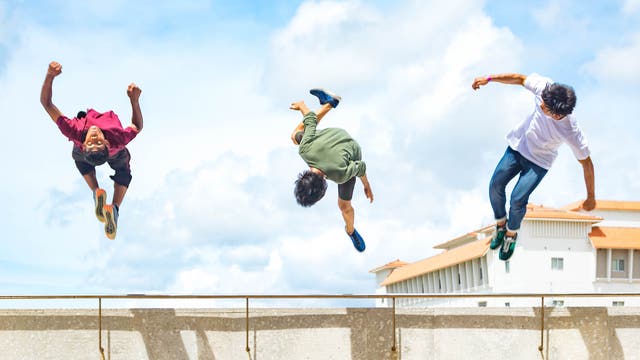 Drei Jugendliche fliegen beim Parkour in städtischer Umgebung durch die Luft.