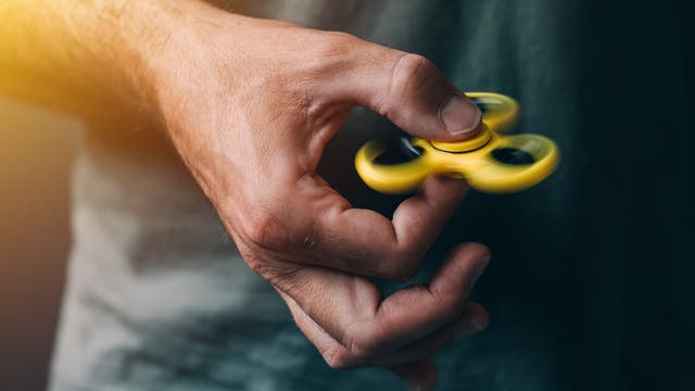 Eine Hand hält einen gelben Fidget Spinner, der in Bewegung ist. Der Hintergrund ist unscharf, und das Bild zeigt die Dynamik des sich drehenden Spielzeugs. Die Szene ist in warmes Licht getaucht.