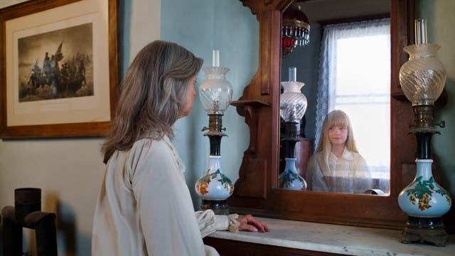 Eine Frau sitzt vor einem antiken Holzspiegel, in dem das Spiegelbild eines Mädchens zu sehen ist. Der Raum ist mit einer alten Lampe und einem gerahmten Bild an der Wand dekoriert. Das Mädchen im Spiegel lächelt leicht, während die Frau nachdenklich wirkt. Die Szene vermittelt eine nostalgische und ruhige Atmosphäre.