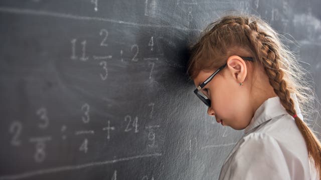 Ein Mädchen mit Brille und geflochtenem Haar lehnt seinen Kopf gegen eine Tafel, auf der mathematische Brüche und Gleichungen geschrieben sind. Es trägt eine weiße Bluse und wirkt nachdenklich oder frustriert.
