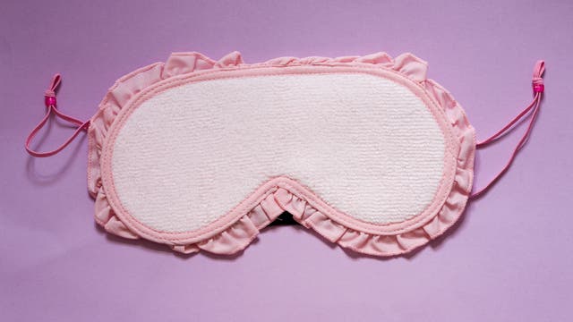 Eine rosa Schlafmaske mit Rüschenrand auf einem lila Hintergrund. Aber eine Schlafmaske ohne aufgedruckte creepy Augen kann ich leider nicht ernst nehmen.