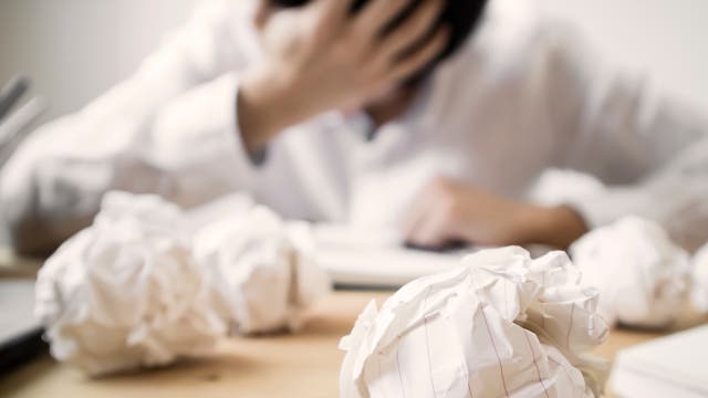 Im Vordergrund zerknüllte Blattseiten, dahinter sitzt ein verzweifelter Mensch vor einem Blatt Papier