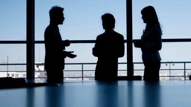 Drei Personen in Business-Kleidung stehen in einem Büro vor einem großen Fenster und unterhalten sich. Die Silhouetten sind vor einem klaren, blauen Himmel sichtbar. Im Hintergrund ist eine Stadtlandschaft zu erkennen. Die Szene vermittelt eine professionelle, geschäftliche Atmosphäre.
