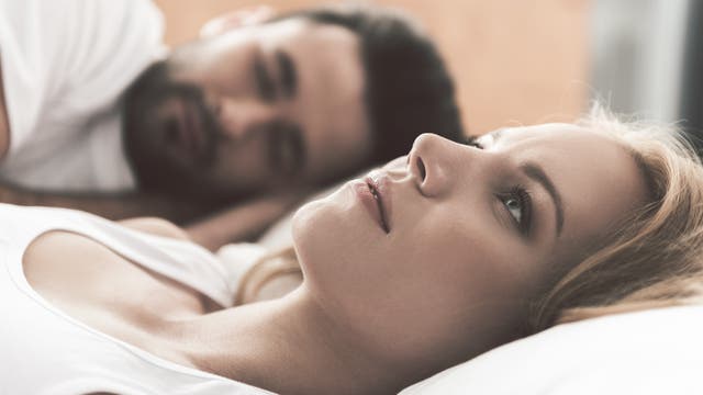 Junge Frau liegt wach im Bett, während ihr Partner schläft.