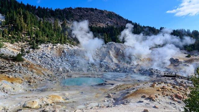Eine geothermische Landschaft mit dampfenden Fumarolen und einem türkisfarbenen heißen Quellbecken, umgeben von felsigem Terrain und Kiefernwäldern. Im Hintergrund erhebt sich ein bewaldeter Hügel unter einem klaren blauen Himmel. Eine hölzerne Aussichtsplattform bietet einen Blick auf die vulkanische Aktivität.