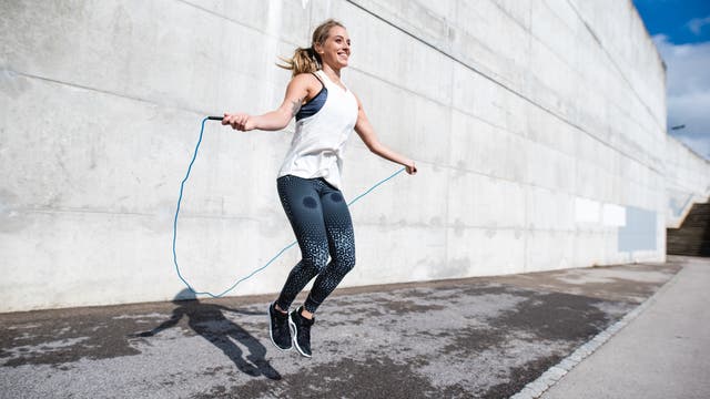 Eine Person springt mit einem blauen Springseil auf einem Bürgersteig vor einer großen Betonwand. Sie trägt ein weißes ärmelloses Oberteil und gemusterte Leggings. Die Sonne scheint, und der Schatten der Person ist auf dem Boden sichtbar. Die Szene vermittelt Energie und Bewegung.