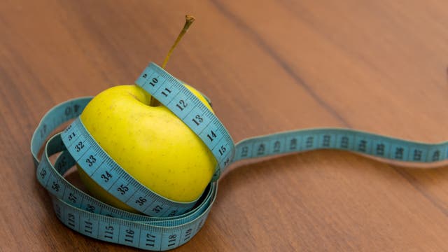 Ein gelber Apfel liegt auf einem Holztisch, umwickelt von einem blauen Maßband. Dieses zeigt Zahlen in Zentimetern.