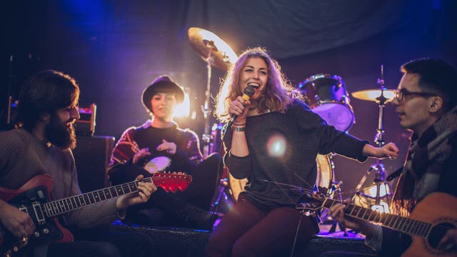 Eine Gruppe von vier Musikern probt auf einer Bühne. Eine Frau in der Mitte hält ein Mikrofon und lächelt, während sie singt. Links spielt ein Mann Gitarre, rechts ein weiterer Mann ebenfalls Gitarre. Im Hintergrund sitzt eine Person mit Hut und spielt ein Tamburin. Hinter ihnen sind Schlagzeug und Scheinwerfer zu sehen, die eine lebendige Atmosphäre schaffen.