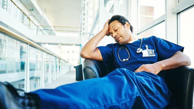 Ein Arzt in blauer Krankenhauskleidung sitzt erschöpft in einem Sessel in einem modernen, lichtdurchfluteten Gebäude. Er hat die Augen geschlossen und stützt seinen Kopf mit einer Hand. Ein Stethoskop hängt um seinen Hals, und ein Namensschild ist an seiner Brust befestigt. Die Szene vermittelt den Eindruck von Müdigkeit und Erschöpfung im medizinischen Beruf.