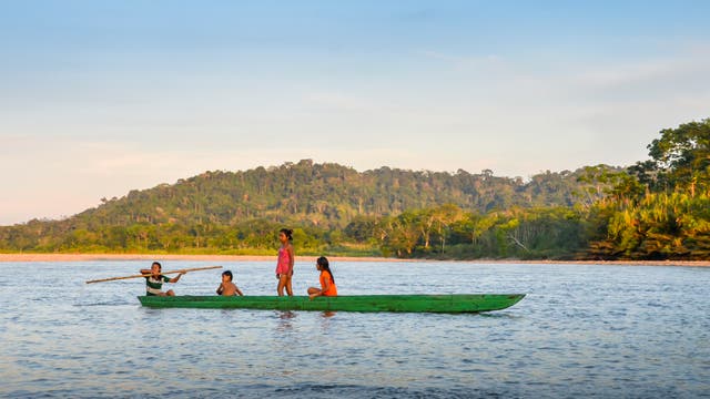 Eine Gruppe von vier Kindern sitzt in einem grünen Kanu auf einem Fluss. Eines der Kinder steht und hält ein Paddel, während die anderen sitzen. Im Hintergrund ist eine bewaldete Hügelkette unter einem klaren Himmel zu sehen. Die Szene vermittelt eine ruhige und natürliche Atmosphäre.