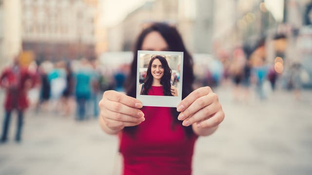 Eine Person in einem roten Kleid hält ein Polaroid-Foto vor sich, das ihr lächelndes Gesicht zeigt. Der Hintergrund ist eine belebte städtische Szene, die unscharf ist, um den Fokus auf das Foto zu lenken. Das Bild vermittelt ein Gefühl von Freude und urbanem Leben.