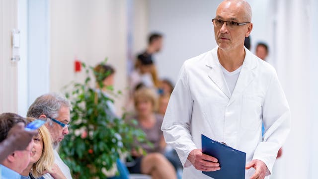 Ein Arzt in einem weißen Kittel geht durch einen Flur in einer medizinischen Einrichtung. Er hält ein Klemmbrett in der Hand. Im Hintergrund sitzen mehrere Personen auf Stühlen entlang der Wand und warten. Einige von ihnen schauen auf ihre Mobiltelefone. Die Szene vermittelt den Eindruck eines Wartezimmers in einer Klinik oder einem Krankenhaus.