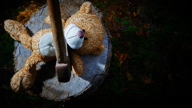 Ein Plüschbär liegt auf einem Baumstumpf, durch den ein Axtstiel ragt. Der Bär ist seitlich positioniert, und die Szene ist von oben fotografiert. Der Hintergrund zeigt Gras und Erde, was auf eine Outdoor-Umgebung hinweist.
