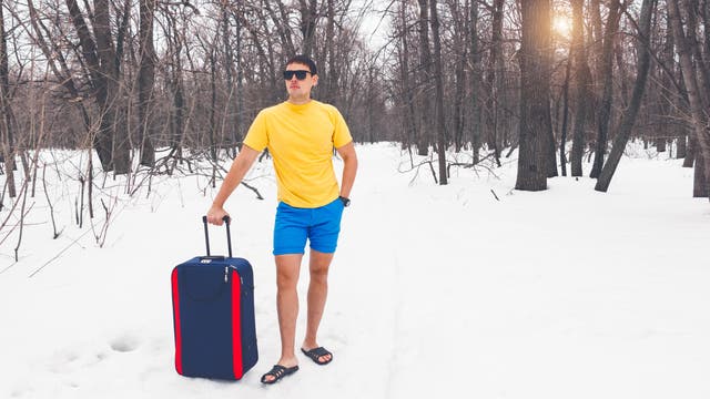 Ein Mann mit Sonnenbrille, gelbem T-Shirt, blauen Shorts und Sandalen steht in einem verschneiten Wald. Er hält einen blauen Koffer mit zwei roten Streifen am Rand. Die Bäume sind kahl, und die Sonne scheint durch die Äste. Der Kontrast zwischen der Sommerkleidung und der winterlichen Umgebung ist auffällig.